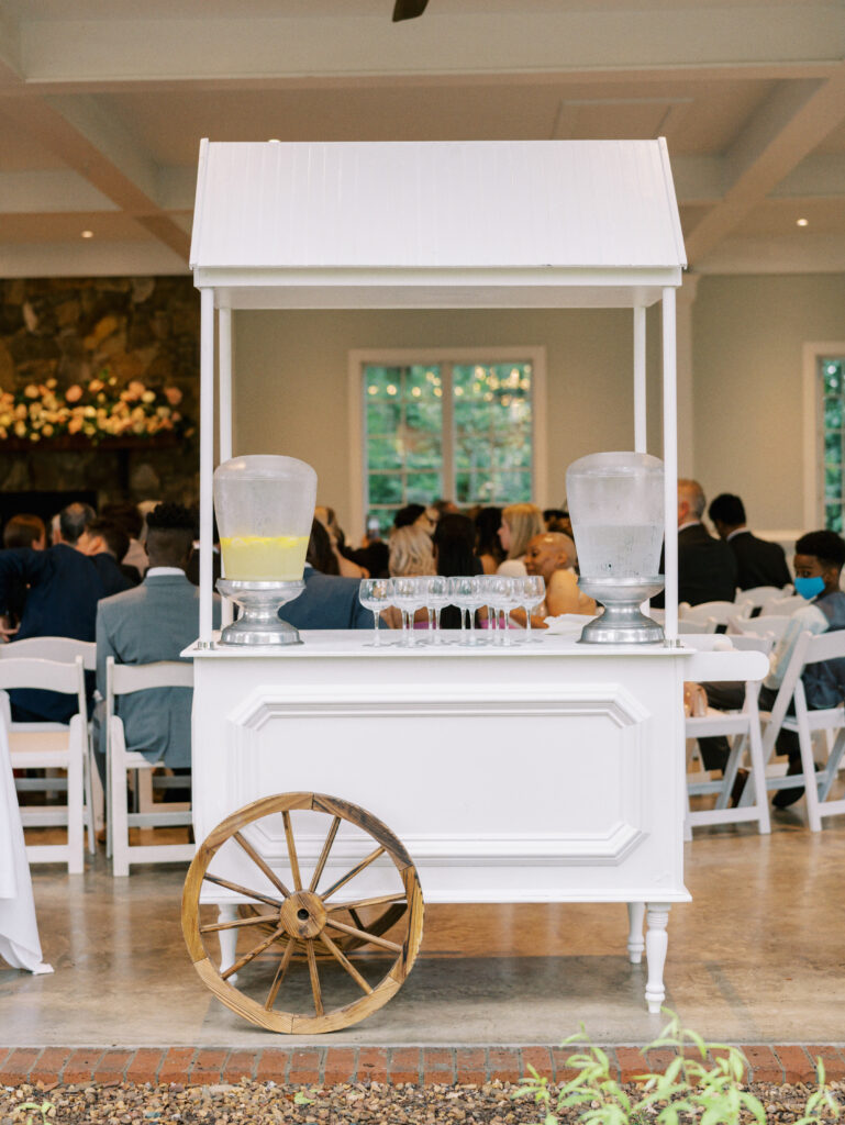 A white drink station with glass dispensers of water and lemonade stands in front of rows of seated wedding guests inside a bright venue. A decorative wooden wheel is attached to the cart.