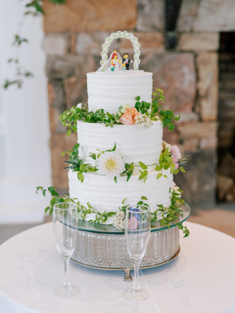 Three-tiered white wedding cake decorated with green leaves and pastel flowers, topped with LEGO bride and groom figures, placed on a silver stand. Two empty champagne flutes sit in front of the cake.