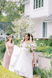 A smiling bride in a white dress holds her bouquet and gown while walking outside with two bridesmaids in pink dresses near a white house and blooming greenery.