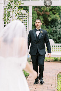 A groom in a black tuxedo smiles as he sees his bride, who is walking toward him in a white dress and veil, outdoors on a brick path with greenery and a white arbor in the background.