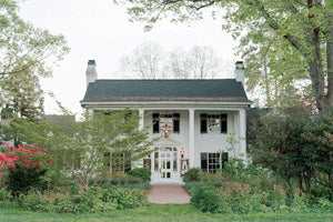 A white two-story house with black shutters and columns, surrounded by green trees and bushes, with a brick walkway leading to the front door.