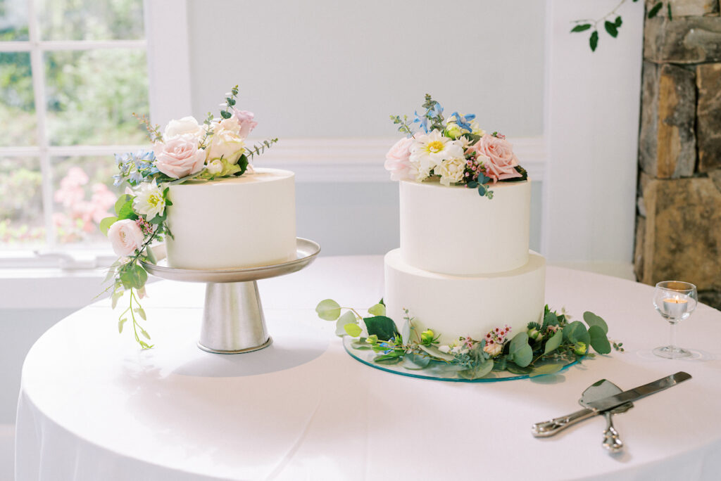 Two elegant white wedding cakes with floral decorations sit on a round table; one cake is single-tiered on a stand, the other is two-tiered with greenery and flowers at the base. A cake knife and server are nearby.