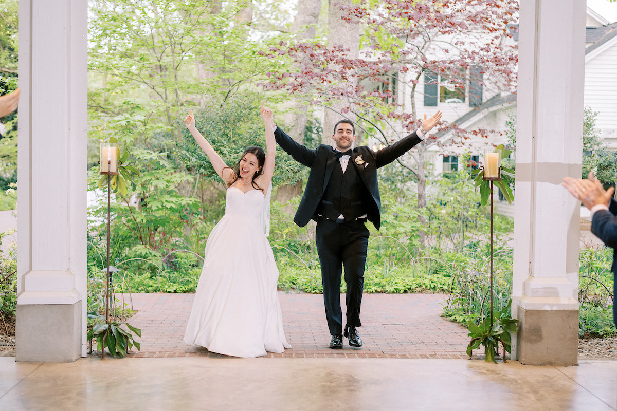 A joyful bride in a white dress and groom in a black tuxedo enter an outdoor venue with arms raised, smiling and celebrating, surrounded by greenery and candle decorations.
