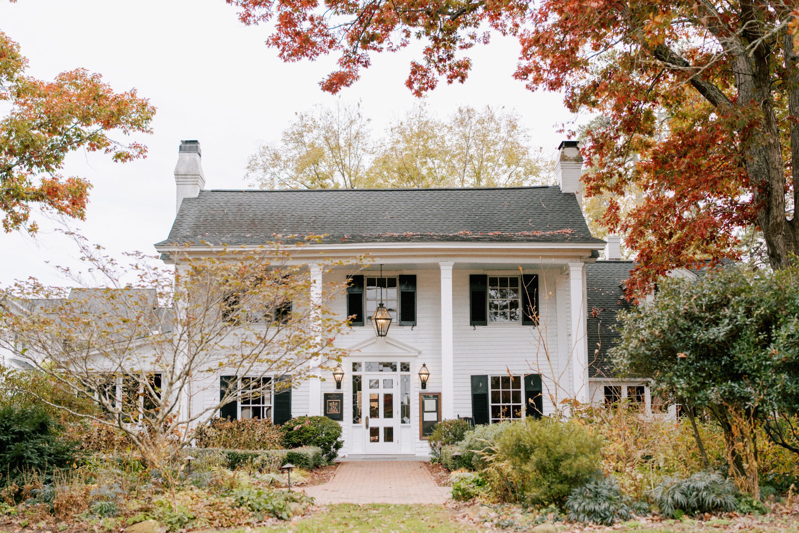 A white two-story house with black shutters and columns, surrounded by autumn trees and shrubs. The pathway leads to a front door with glass panels and lantern hanging above.