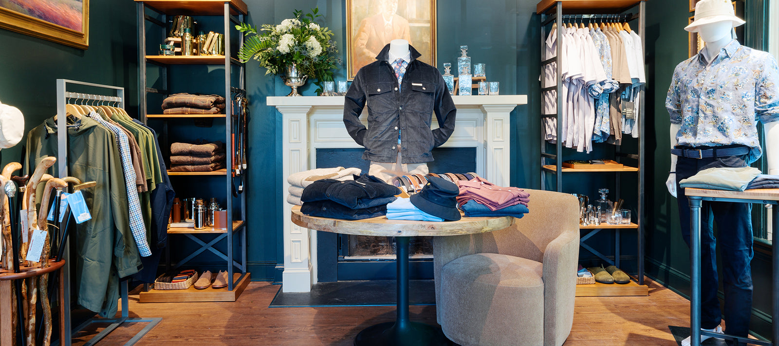 A stylish men’s clothing boutique with folded shirts and pants on a round table, jackets and shirts hanging on racks, and accessories throughout the room, set against elegant green walls and a decorative white fireplace.