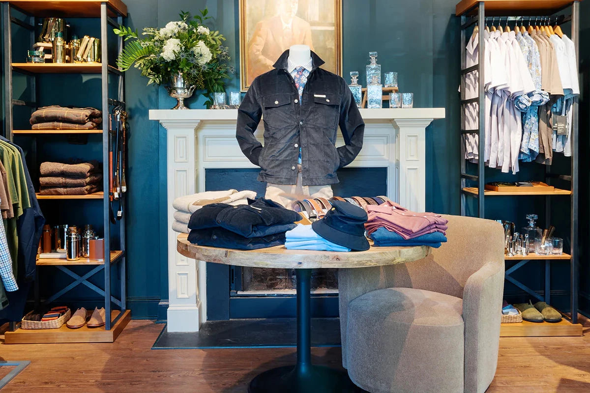 A men’s clothing boutique with folded shirts and sweaters on a round table, a mannequin in a dark jacket, shelves with more clothes, and a fireplace with a portrait above it in the background.
