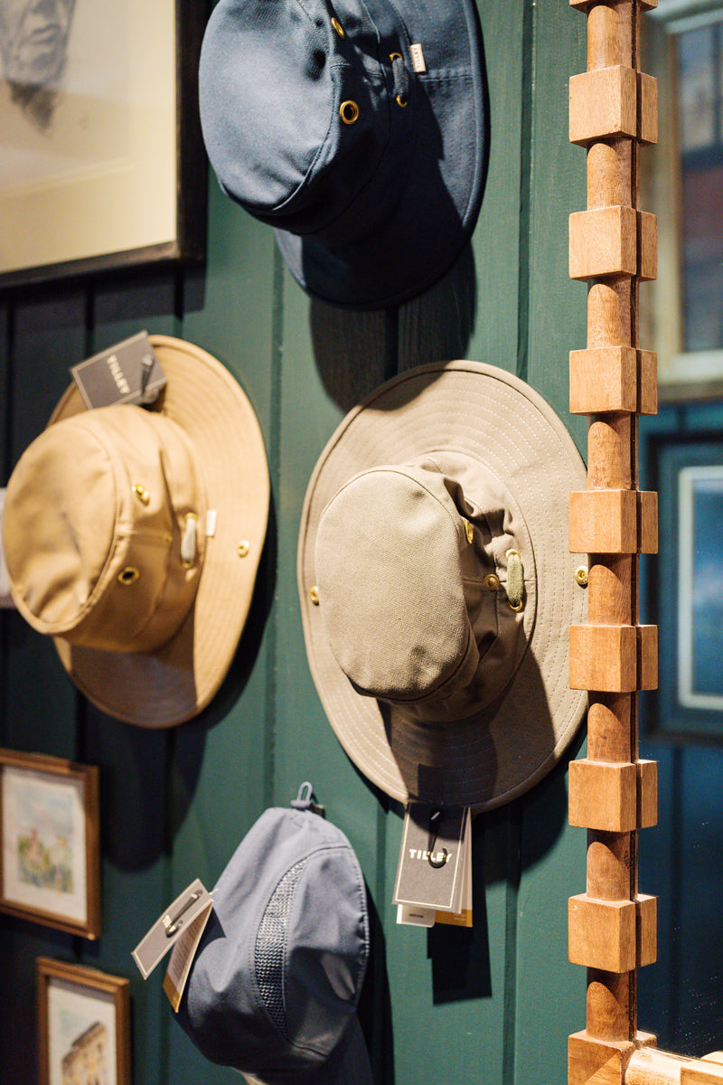 Four wide-brimmed hats in shades of beige, olive, and blue hang on a dark green wooden wall, each with a tag attached. A wooden decorative piece and framed artwork are partially visible nearby.