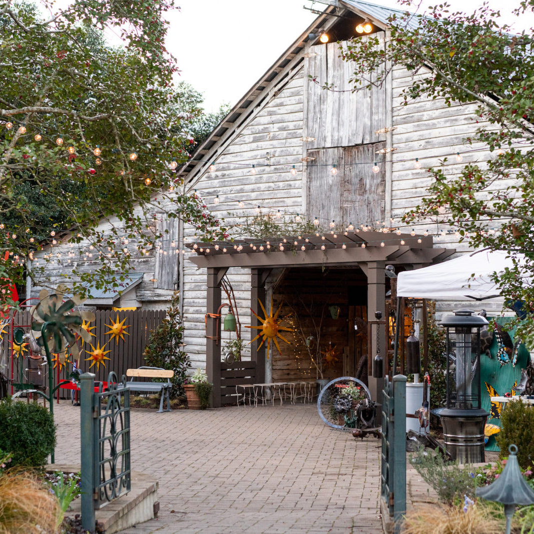 A rustic wooden barn with weathered siding is decorated with string lights and metal star ornaments. Trees and plants line the brick pathway leading to the entrance, creating a cozy outdoor atmosphere.
