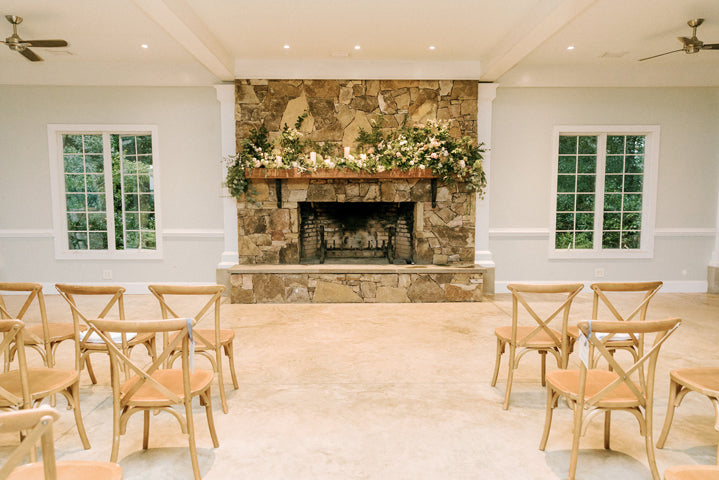 A bright room with wooden chairs arranged in rows facing a large stone fireplace decorated with green foliage and white flowers. Two large windows flank the fireplace, and the floor is light-colored.
