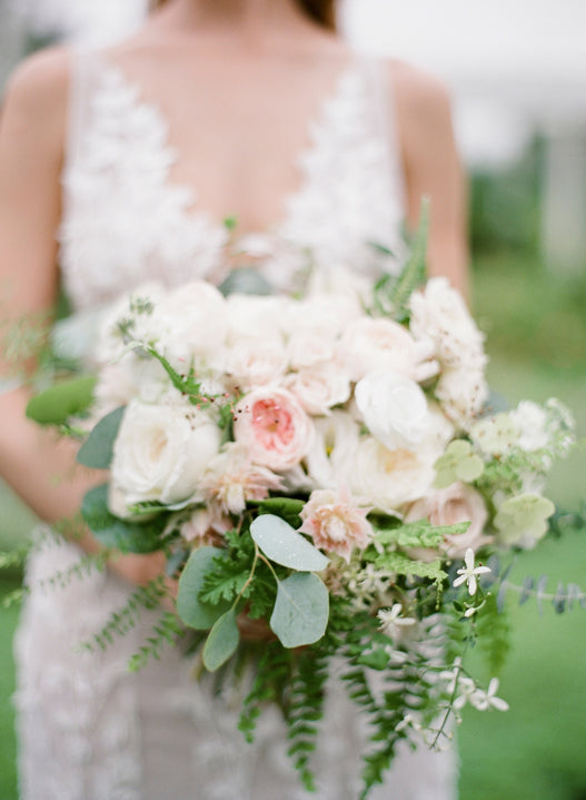 A bride in a white lace dress holds a bouquet of white and blush pink flowers with green foliage, standing outdoors with a blurred natural background.