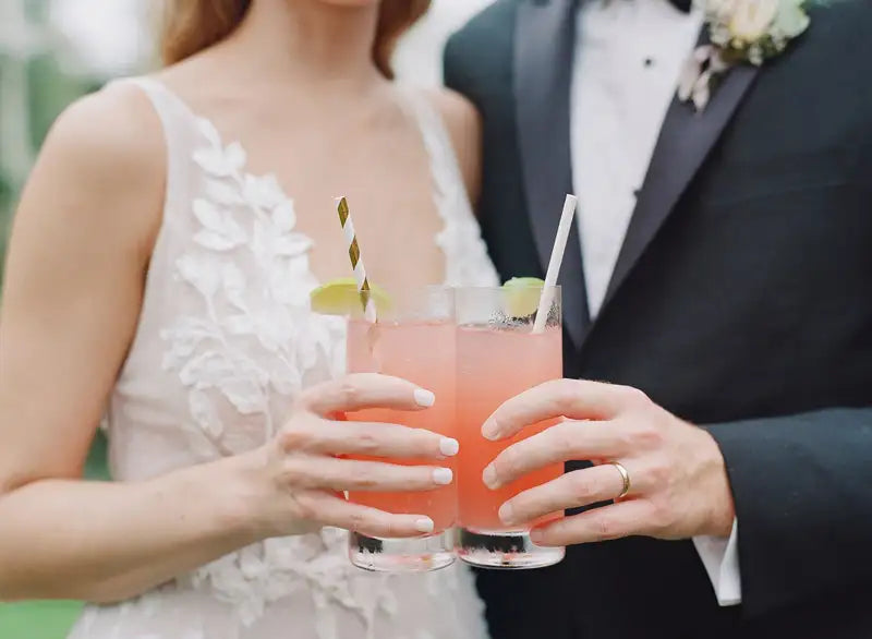 A couple dressed in formal wedding attire holds pink cocktails with lime wedges and striped straws, standing close together and smiling, with their faces partially out of frame.