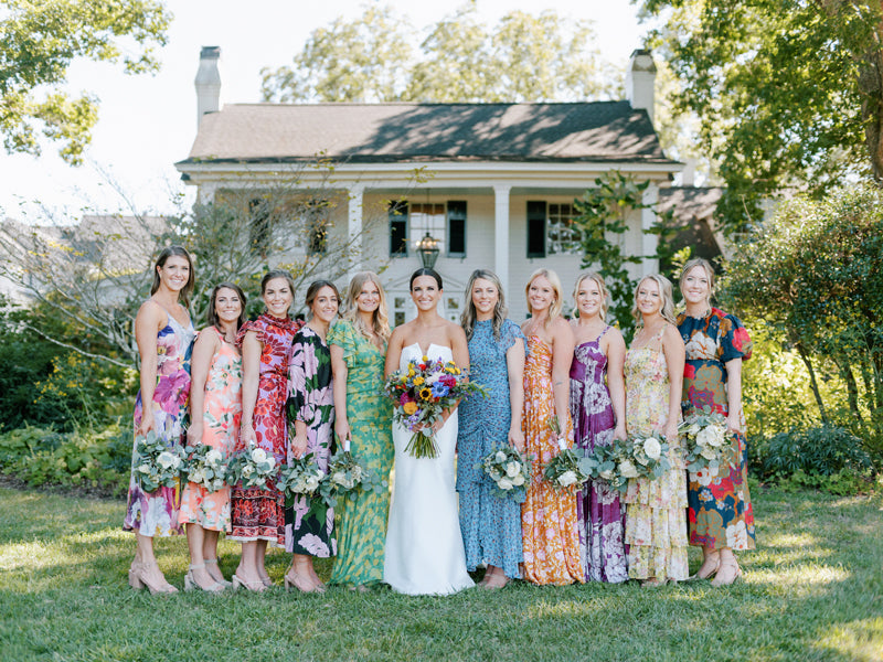 A bride in a white dress stands with eleven bridesmaids in colorful, patterned dresses, holding bouquets, outside in front of a large, white house surrounded by greenery on a sunny day.