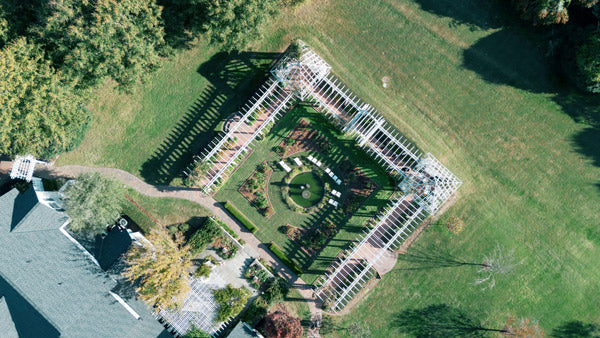 Aerial view of a square garden with a circular lawn at its center, surrounded by a white pergola, pathways, and greenery, set in a large grassy area with scattered trees.