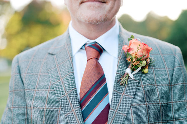 A close-up of a man in a plaid suit jacket, white shirt, and striped tie, wearing a boutonniere with orange and pink flowers pinned to his lapel. The background is blurred greenery.