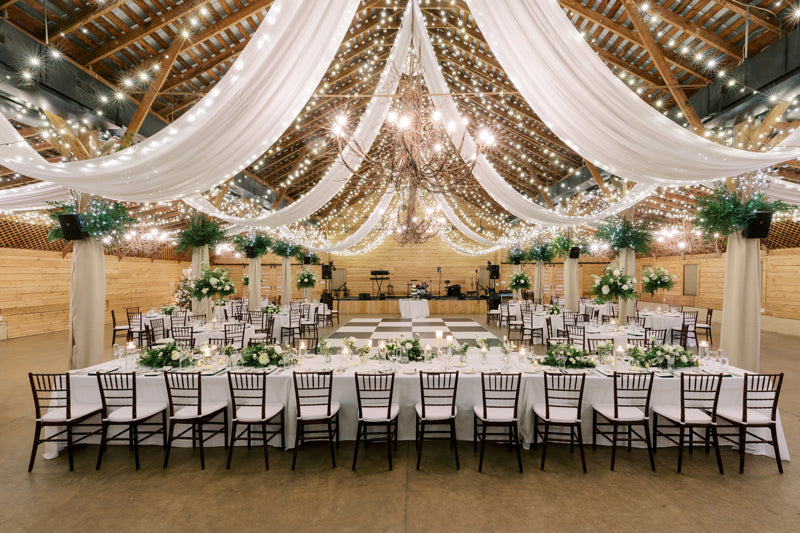 A beautifully decorated barn wedding reception with draped white fabric, string lights, elegant floral centerpieces, and tables arranged in a U-shape around a dance floor.