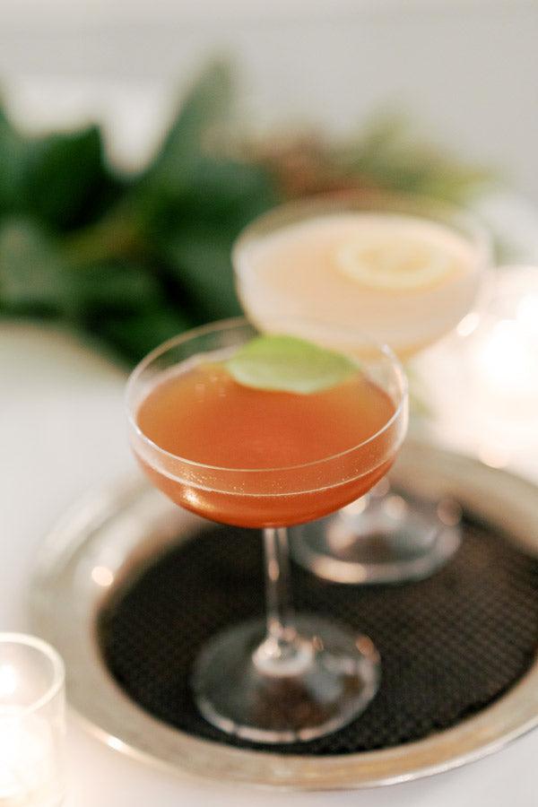 Two elegant cocktails in coupe glasses sit on a silver tray; one drink is amber with a lime garnish, the other is pale with a lemon slice. A blurred green background and candles add a soft, inviting ambiance.