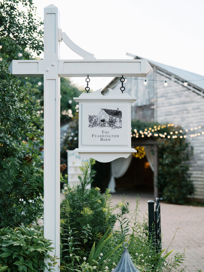 A white hanging sign for “The Fearrington Barn” stands among greenery, with a rustic barn and string lights visible in the softly focused background.