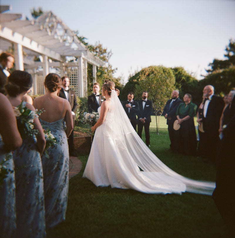A bride in a flowing white gown and veil stands outdoors at an altar, facing her groom. Bridesmaids, groomsmen, and guests in formal attire surround the couple under a pergola on a sunny day.