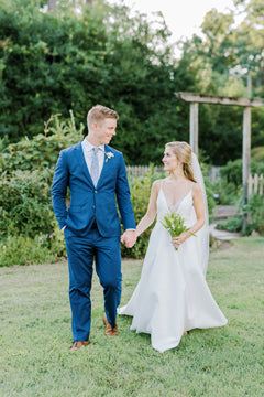 A bride in a white dress and a groom in a blue suit walk hand-in-hand on a grassy lawn, smiling at each other, with greenery and a wooden garden arbor in the background.
