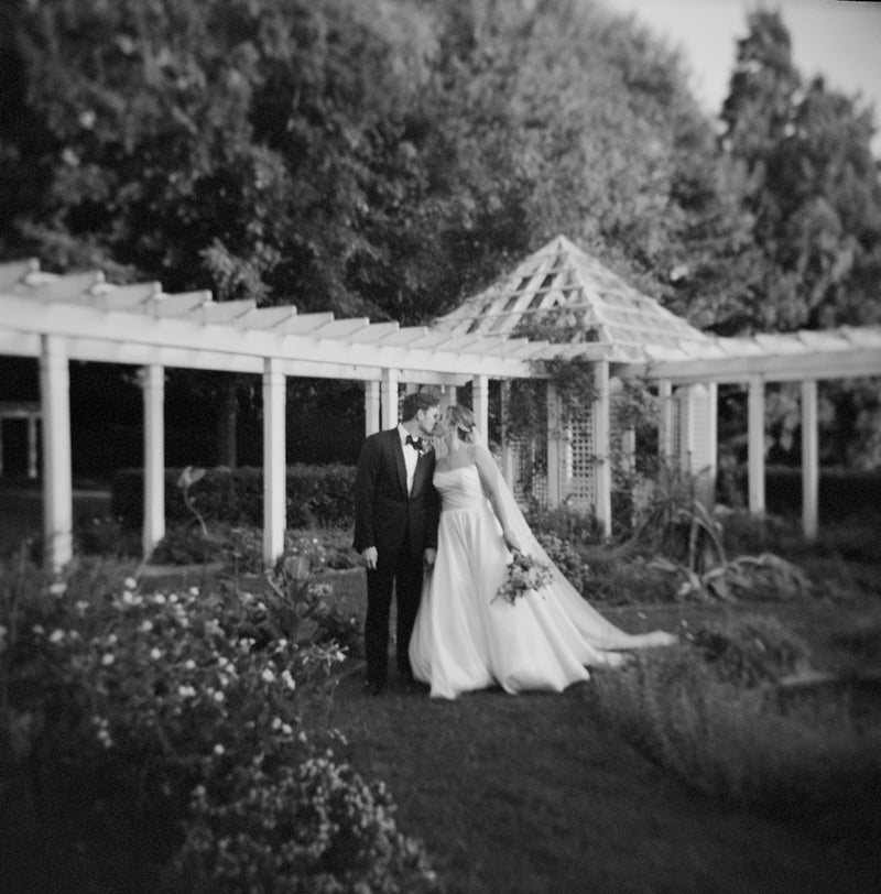 A black-and-white photo of a bride and groom standing close together in a garden, under a white pergola. The bride holds a bouquet and wears a long dress, while the groom is in a suit. The image has a soft, dreamy focus.