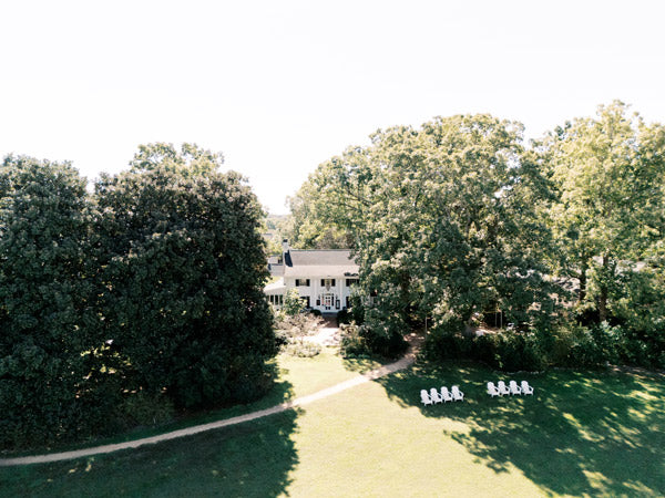 A large white house is partially hidden among tall green trees, with a grassy lawn in front. Several white chairs are arranged in rows on the lawn near a dirt path under a sunny sky.