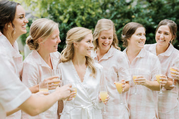 A group of women in matching light pajamas, holding glasses of orange juice, smile and laugh together outdoors with greenery in the background. One woman in a white robe stands in the center.