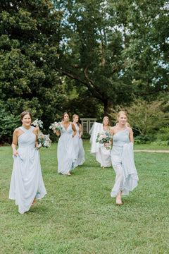 Five women in light blue dresses walk barefoot on grass, holding bouquets of flowers. They are smiling and appear to be bridesmaids in an outdoor garden setting with trees in the background.
