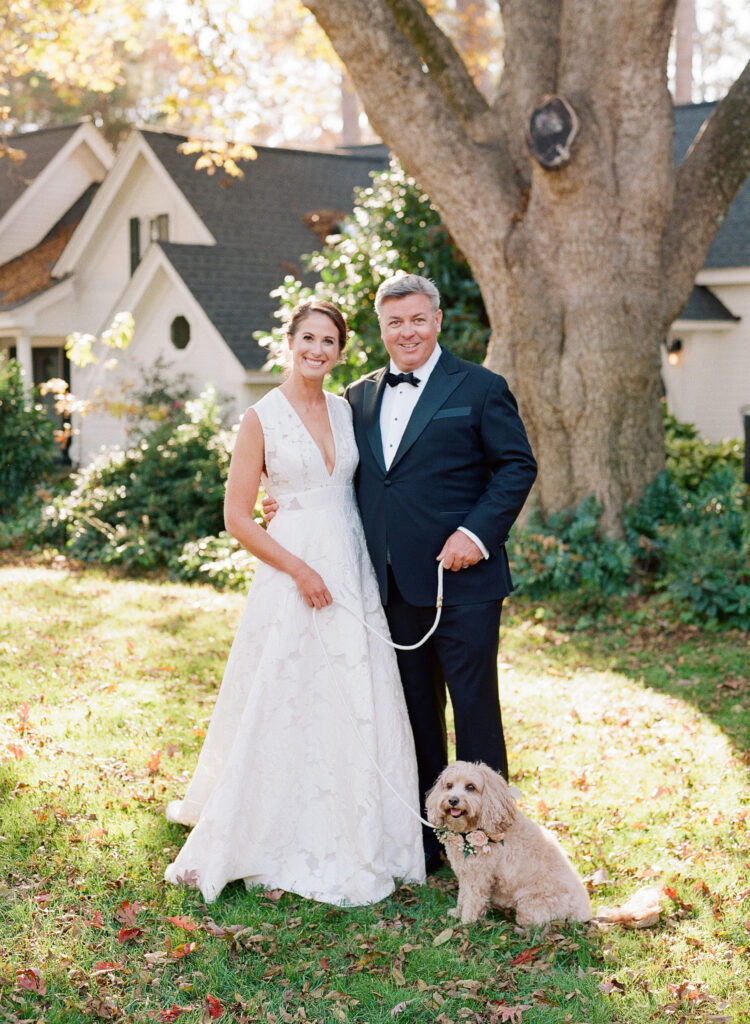 A smiling bride and groom stand outdoors in front of a large tree, holding a small, fluffy dog on a leash. The bride wears a white gown, and the groom wears a dark suit. Sunlight filters through the trees onto the green lawn.