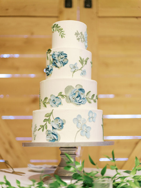 A four-tiered white cake decorated with blue and green floral designs sits on a silver stand, with wooden doors and greenery in the background.