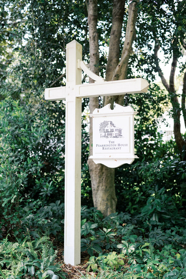 A white wooden signpost stands among green foliage, displaying a hanging sign for 
