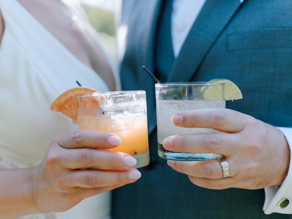 A close-up of a couple in formal attire holding cocktails garnished with citrus slices, toasting together. The woman wears a white dress and the man wears a blue suit with a teal tie.