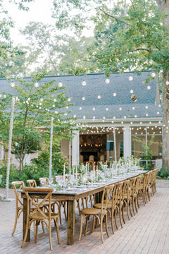 A long wooden table with wooden chairs is set for an outdoor event. The table is decorated with glassware, candles, and greenery. String lights are hung above, and a building with large windows is in the background.