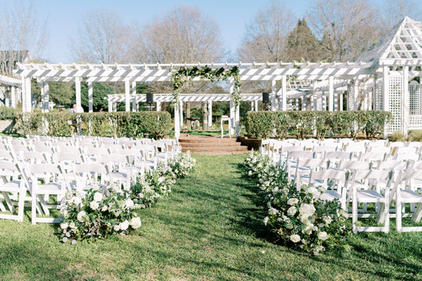 Rows of white chairs arranged on either side of a grassy aisle decorated with white flowers, facing an outdoor pergola altar adorned with greenery and flowers under a clear blue sky.