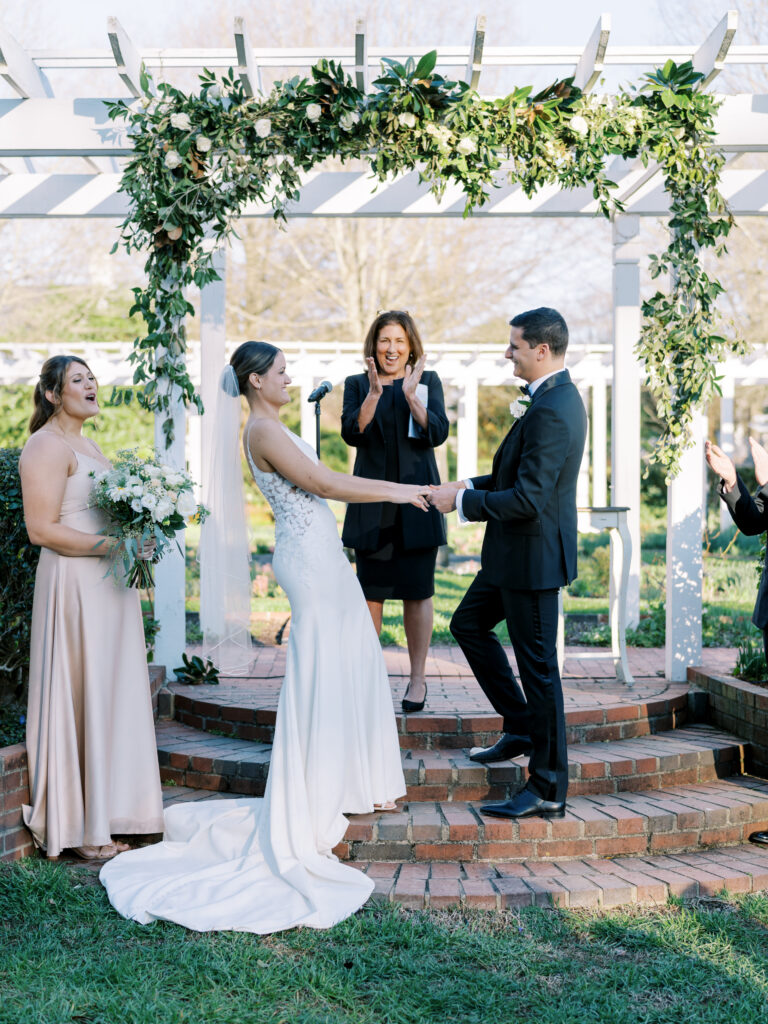 A bride and groom hold hands and smile at each other during an outdoor wedding ceremony. The officiant stands behind them, and a bridesmaid holding a bouquet stands to the side under a floral archway.