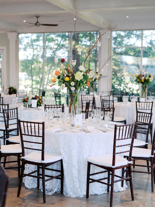 Elegant reception hall with round tables covered in white lace tablecloths, surrounded by dark wooden chairs. Each table features a tall floral centerpiece with greenery and candles. Large windows let in natural light.