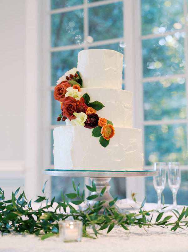 A three-tiered white wedding cake decorated with red, orange, and white flowers, displayed on a silver stand with greenery and a candle on the table, in front of large windows.