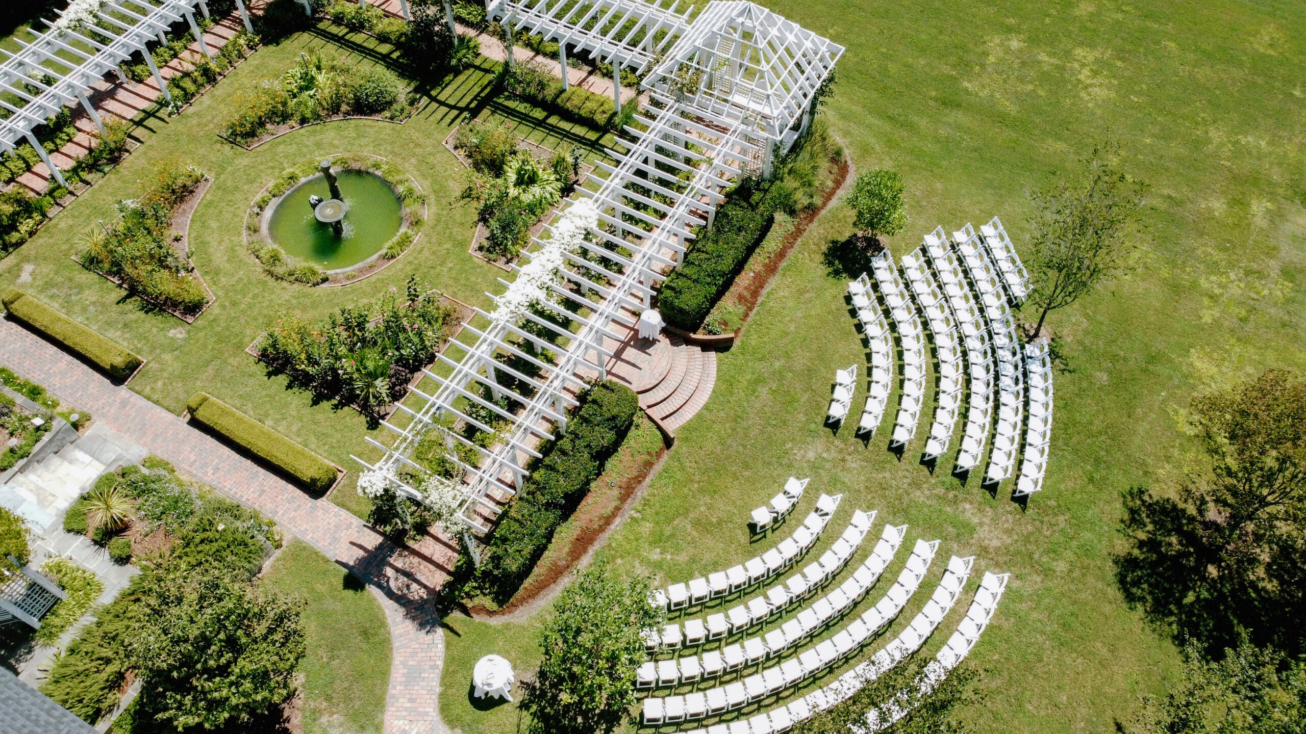Aerial view of an outdoor wedding venue with rows of white chairs facing a pergola and a circular garden fountain, surrounded by green lawns and landscaped pathways.
