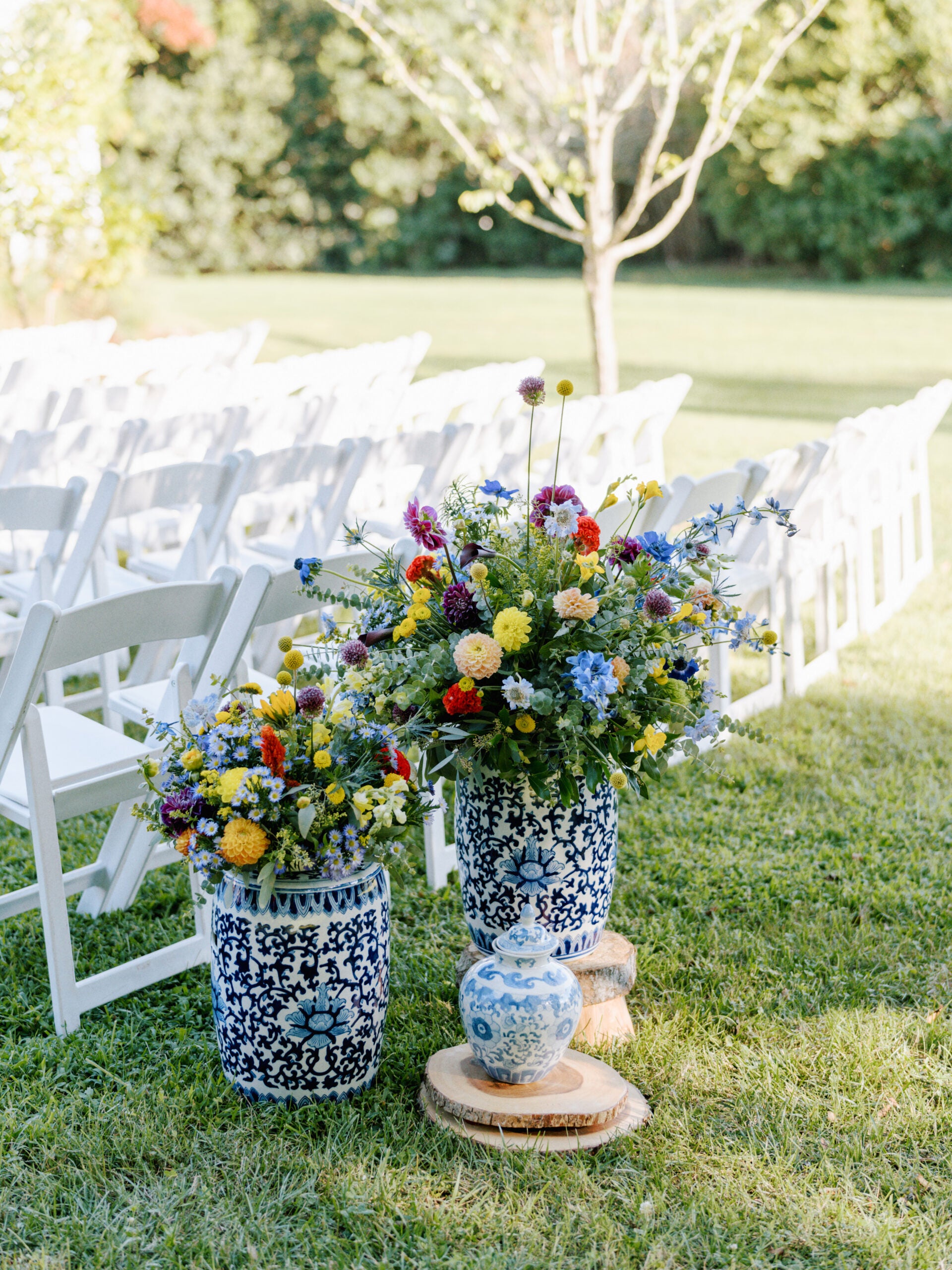 Two blue and white vases filled with colorful flowers sit on wooden slabs beside rows of white folding chairs arranged outdoors on a grassy lawn, with a small matching decorative jar in front.