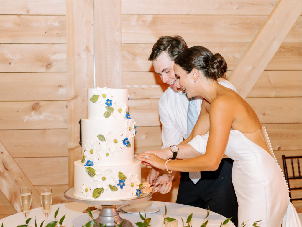 A bride and groom, smiling and dressed in wedding attire, cut a white, three-tiered cake decorated with flowers at their wedding reception in a wooden-paneled venue.