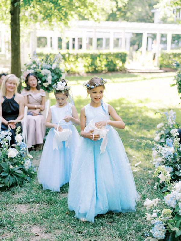 Two young flower girls in light blue tulle dresses and flower crowns walk down an outdoor aisle with white flower petals, surrounded by seated guests and lush greenery on a sunny day.