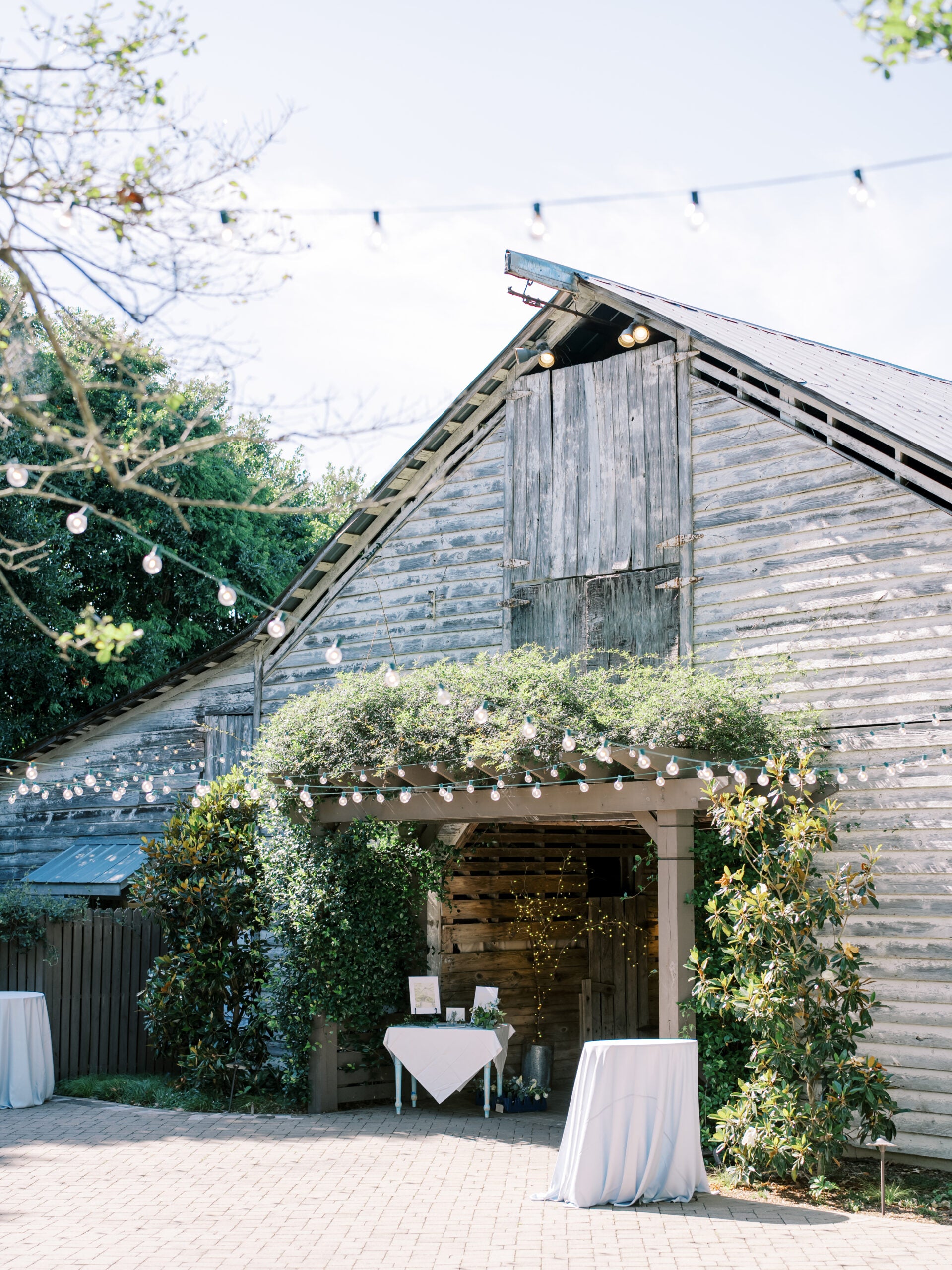 A rustic wooden barn decorated with string lights and greenery, with small round tables covered in white cloths set up outside on a sunny day.
