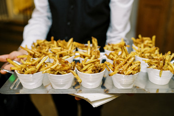 A person holding a tray filled with small white cups, each containing a serving of golden French fries.