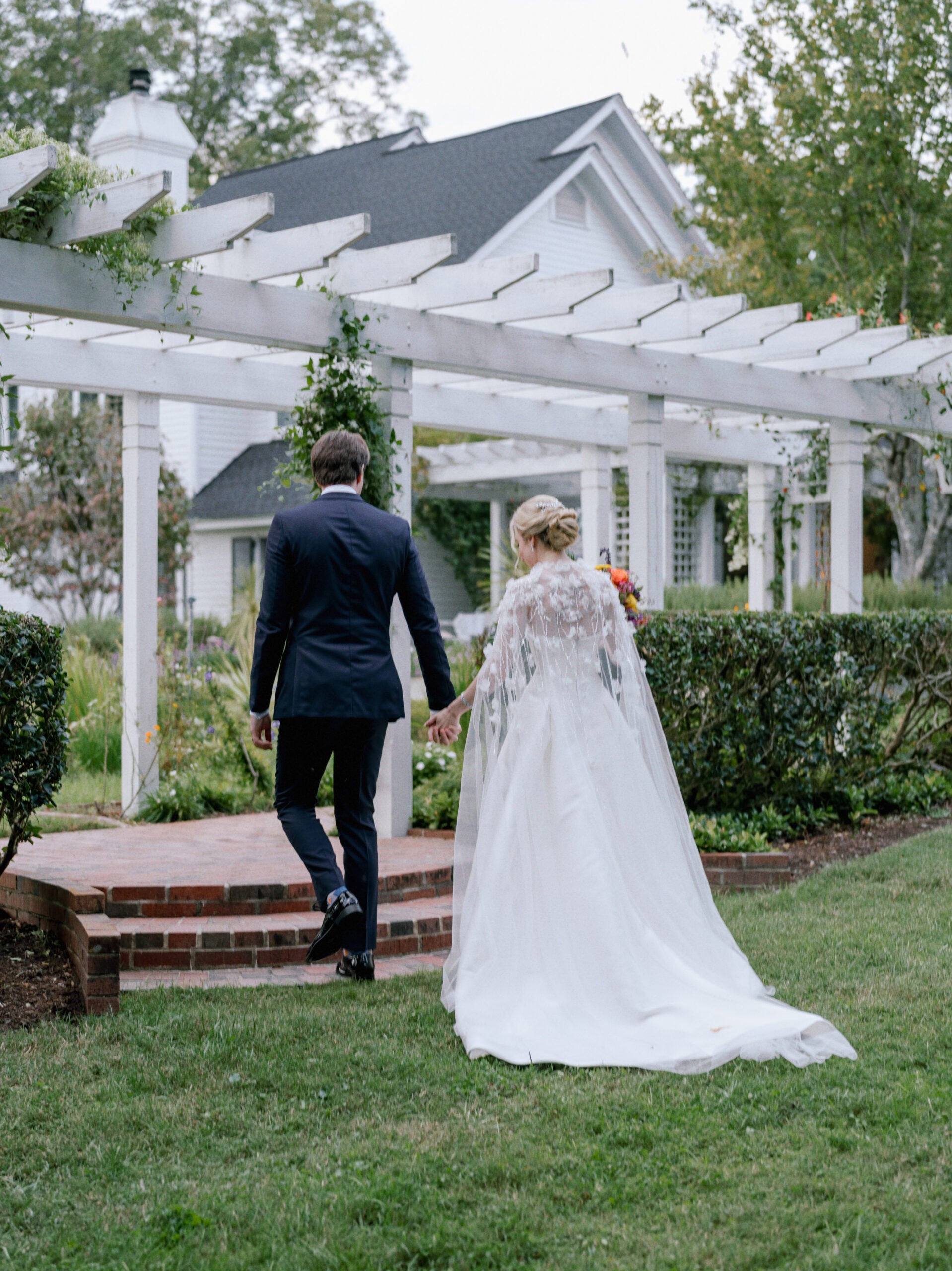 A bride in a long white gown and a groom in a navy suit walk hand in hand under a white pergola in a garden, with a white house in the background.
