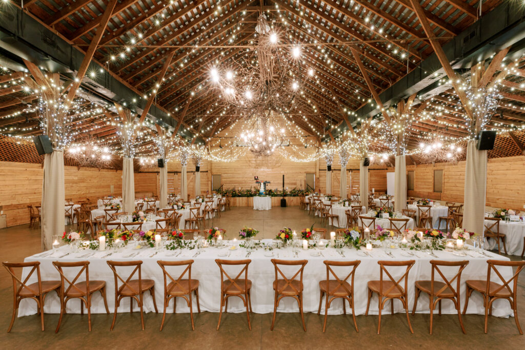 A rustic wedding reception hall with wooden beams, string lights, and chandeliers. Long tables with white tablecloths and floral centerpieces are set with wooden chairs, ready for guests.