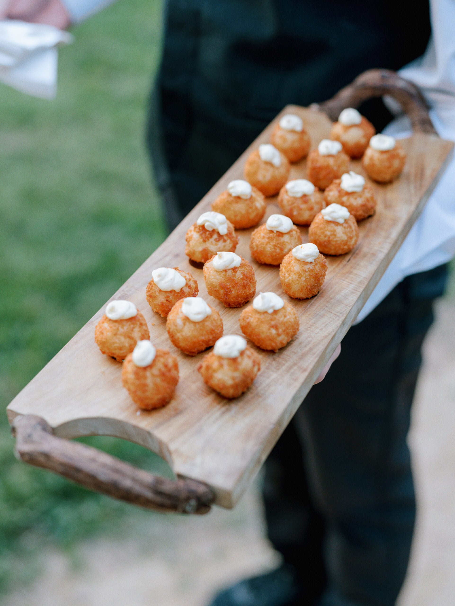 A person holds a wooden serving tray topped with rows of small, golden-brown fried appetizers, each garnished with a dollop of creamy white sauce. The background is outdoors and slightly blurred.