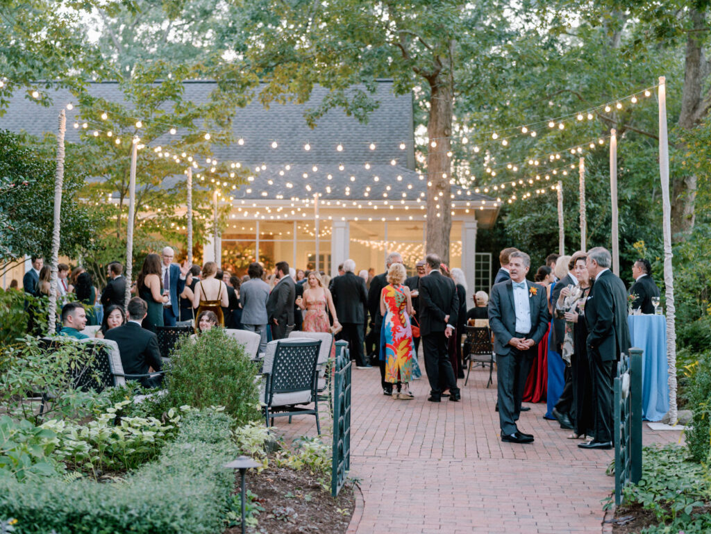 A group of people dressed in formal attire gather outdoors under string lights for an event; some stand and chat on a brick pathway, while others sit among lush greenery near a house.