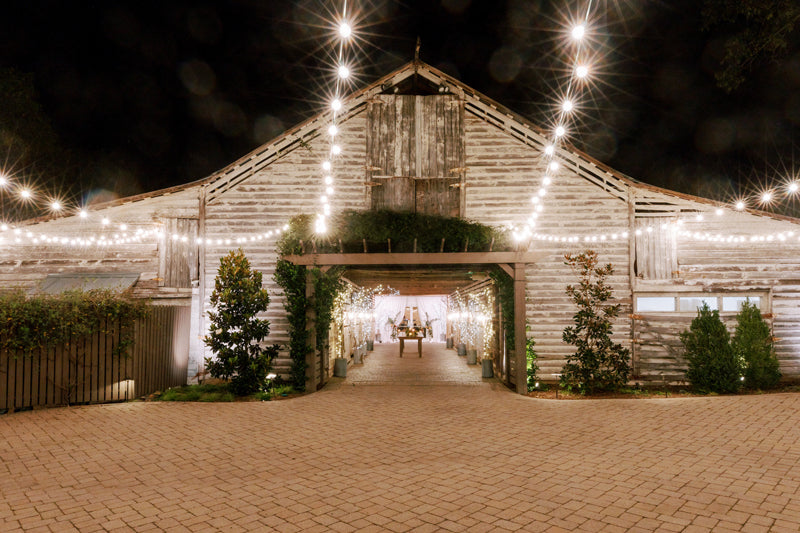 A rustic wooden barn at night is decorated with glowing string lights, creating a warm, inviting atmosphere. The barn entrance is open, revealing a softly lit interior and greenery along the pathway.