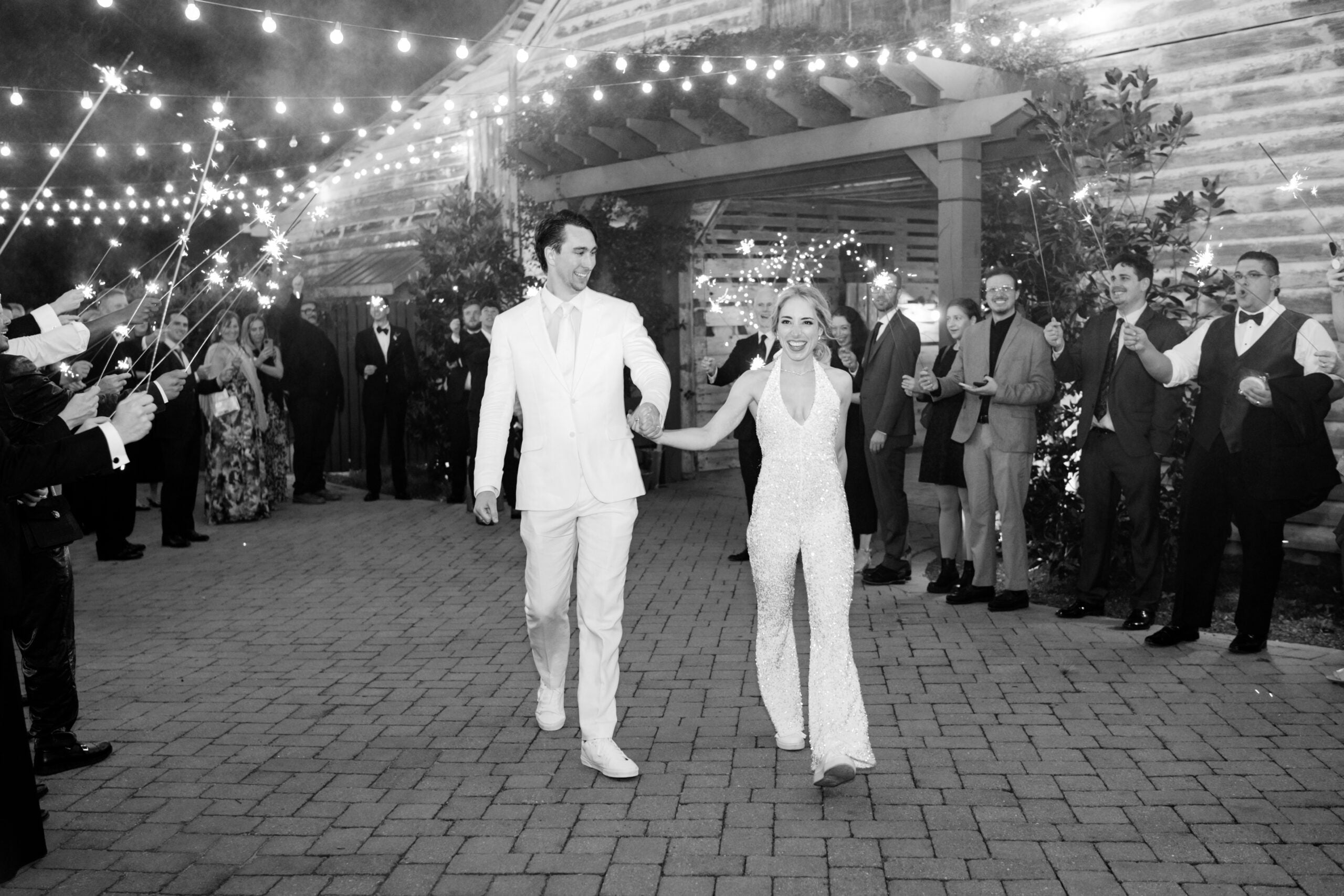 A joyful couple, dressed in white, walk hand in hand under string lights as guests cheer and hold sparklers, celebrating outside at night.