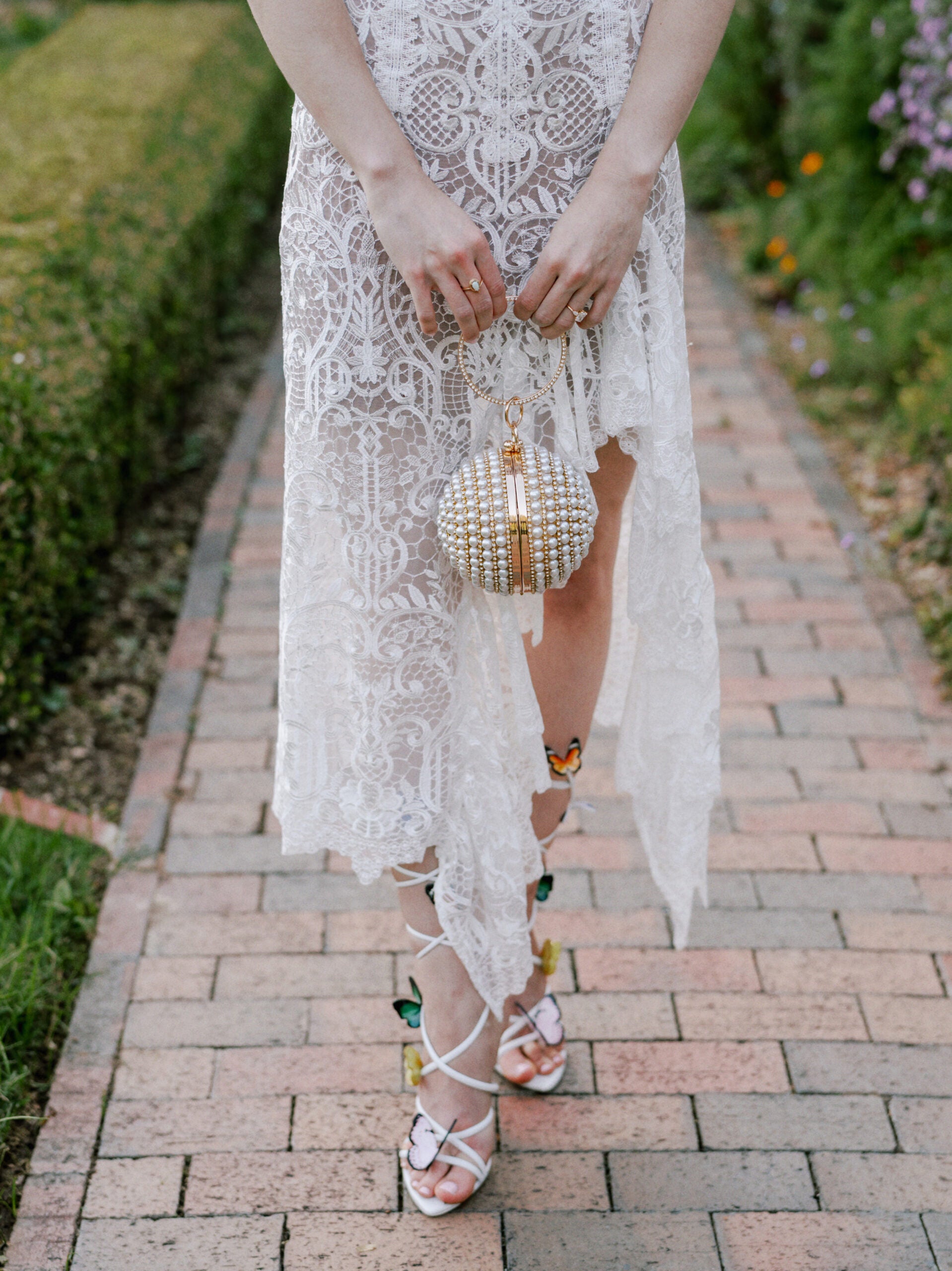 A woman in a white lace dress stands on a brick path, holding a round, beaded handbag. She wears strappy sandals with butterfly details. The garden background is green with flowers. The photo is cropped at her shoulders.