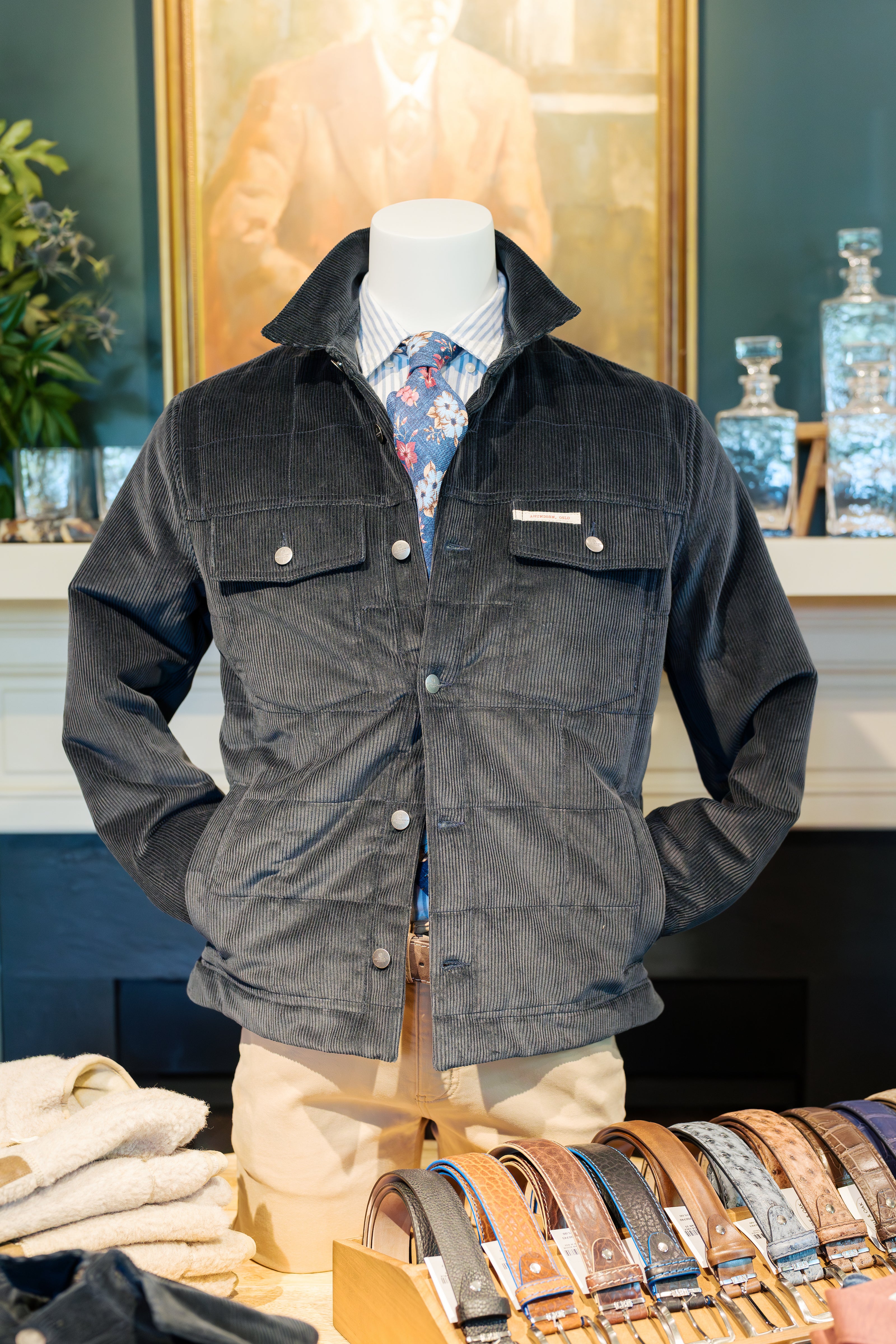 A mannequin dressed in a dark corduroy jacket, light blue striped shirt, and floral tie stands behind a display of assorted leather belts in a clothing store. A blurred portrait painting hangs in the background.